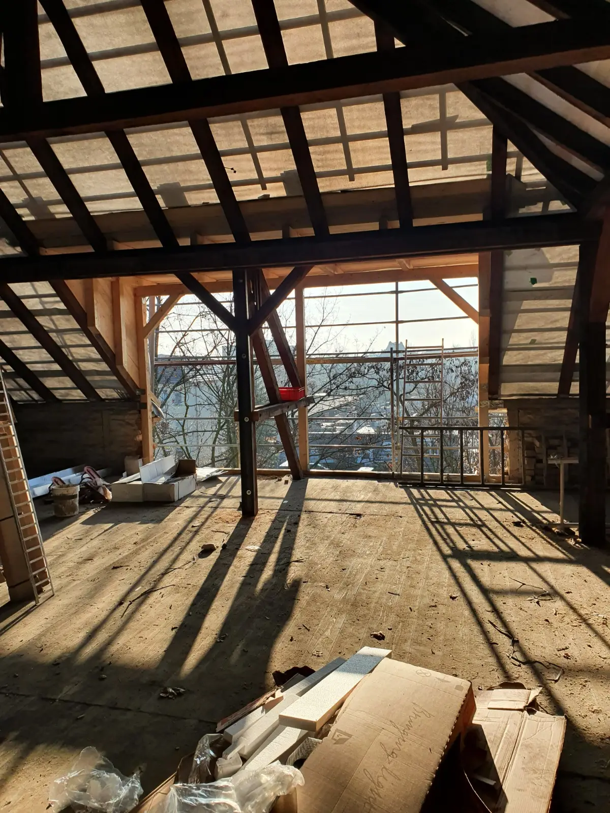 Blick in das Innere eines Dachgeschosses im Rohbauzustand mit sichtbarer Holztragkonstruktion und starkem Sonneneinfall durch die offene Fensterfront.